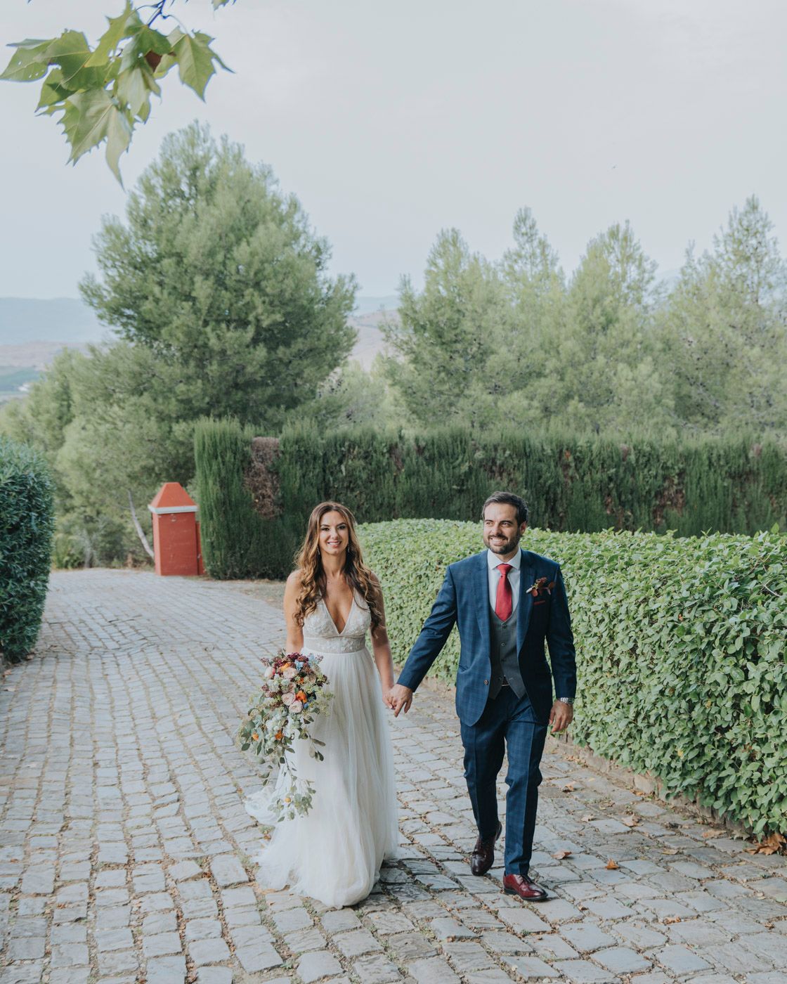 A couple walks hand in hand along a stone pathway surrounded by manicured hedges and trees on their wedding day.
