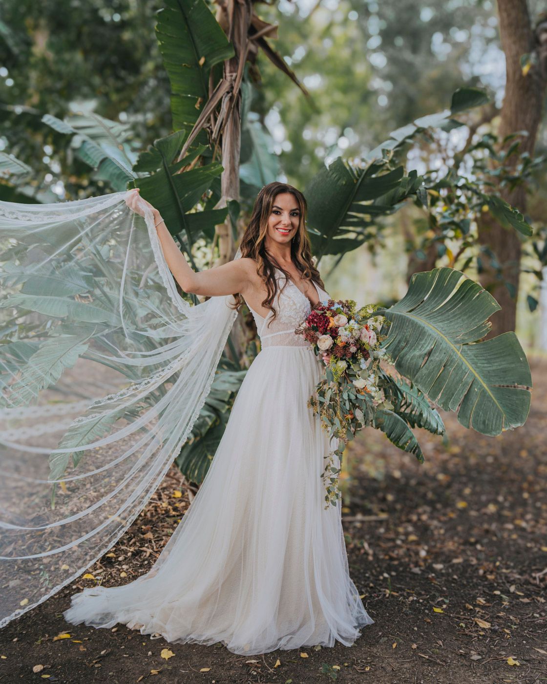 Ethereal bridal photo with flowing veil and garden bouquet amidst lush tropical foliage and natural lighting.