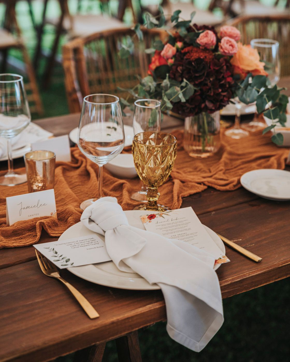 Elegant wedding table setting with terracotta runner, floral centerpiece, gold glassware and white napkins on wooden table.