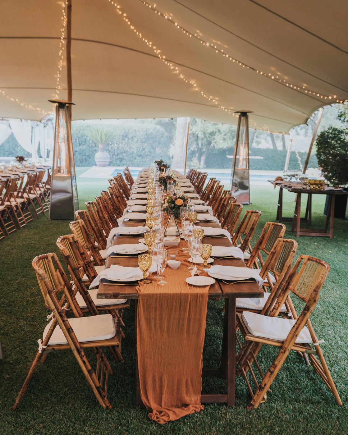 Long banquet table with wooden chairs set under a white tent with string lights and peach table runner for outdoor wedding.