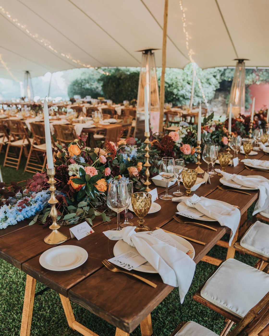 Rustic outdoor wedding reception under white tent with elegant table settings, wooden chairs, and floral centerpieces.