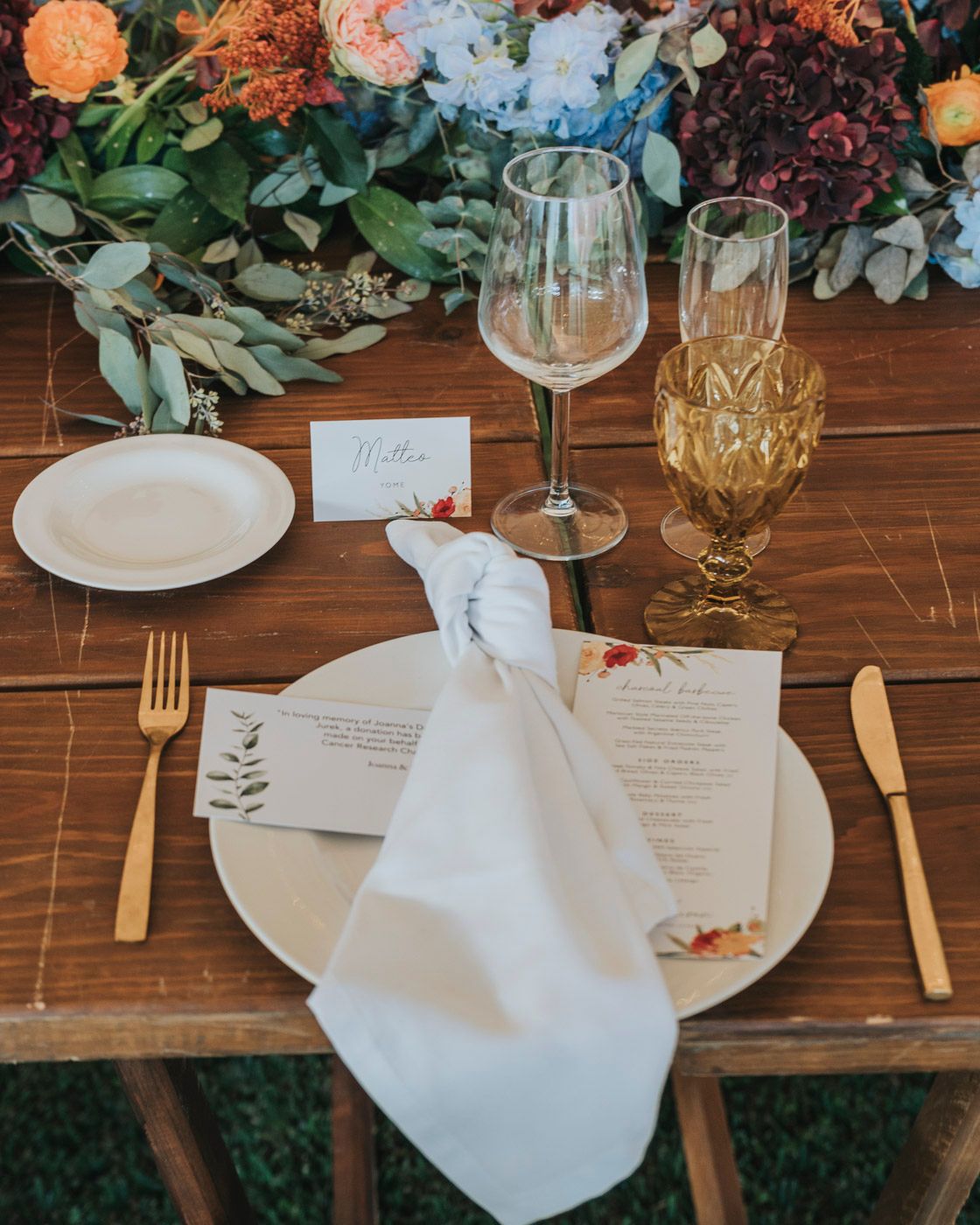 Elegant table setting with wine glasses, menu card, and floral arrangements on wooden surface for wedding reception.