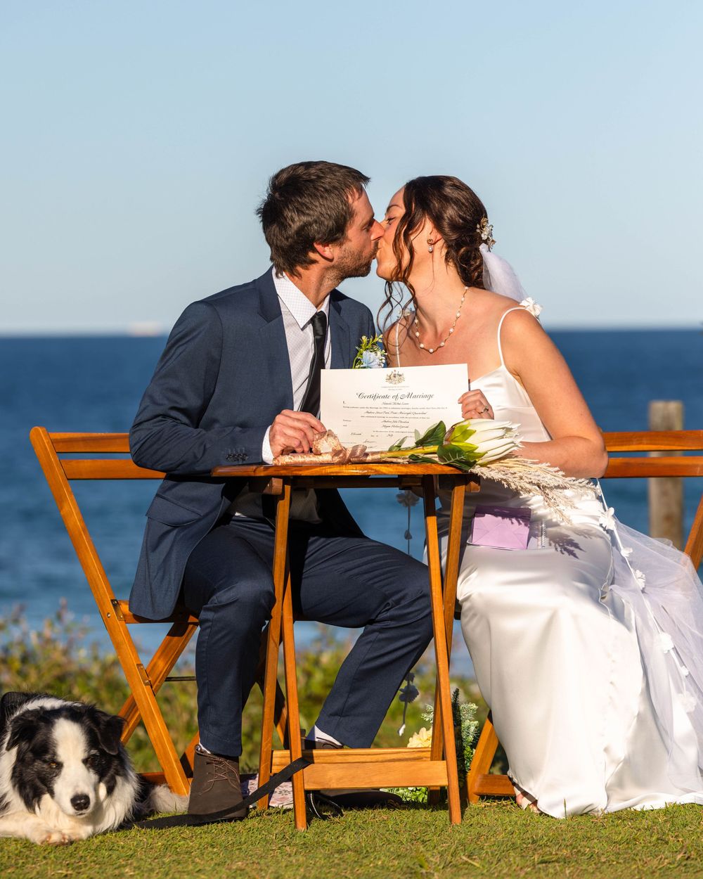 Husband and wife holding up a newly married sign giving each other a kiss with the beautiful blue ocean behind them and their dog over left.