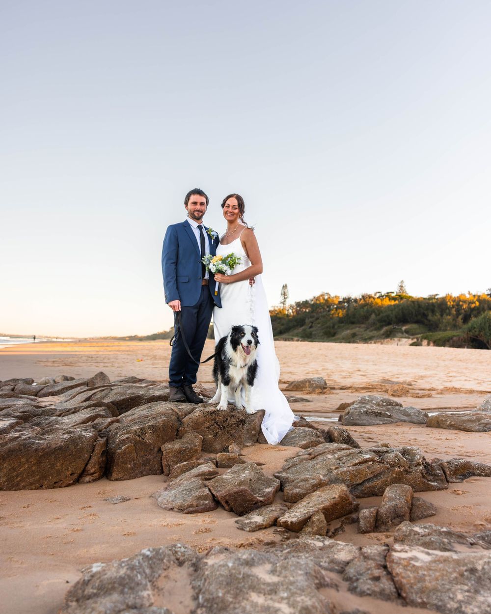 wide image of husband, wife and their dog centre frame standing on the rocks at Coolum Beach Sunshine Coast Queensland.