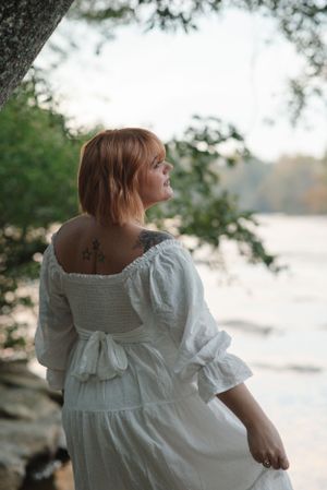 Woman in white dress poses by a lake showcasing a back tattoo and ruffled sleeves.