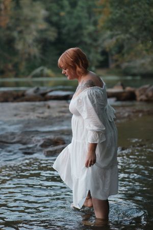 Person in flowing white dress wades in shallow river water surrounded by rocks.
