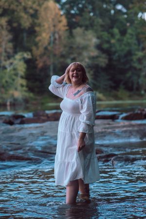 A person in a white dress stands knee-deep in flowing river water at dusk.