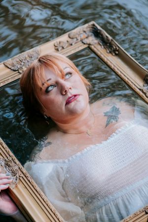 A close-up portrait through an antique frame showing tattoos and a white dress in water.