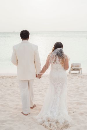 A couple in white wedding attire walks hand in hand on a sandy beach near the ocean.