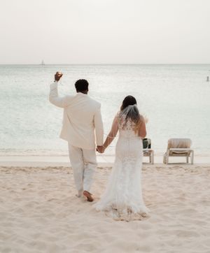 A newlywed couple holds hands while walking on a beach with lounge chairs in the distance.