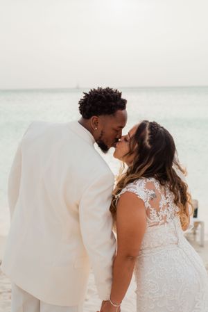 A couple in white wedding attire share a romantic kiss on a sunny beach.