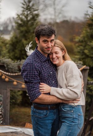 A couple embraces outdoors in casual fall attire with evergreen trees and string lights in the background.