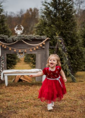 A child in a sparkly red holiday dress twirls and plays in a rustic outdoor setting with evergreen trees and string lights.