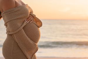 Close-up of pregnant belly wrapped in beige knit dress against sunset ocean backdrop.