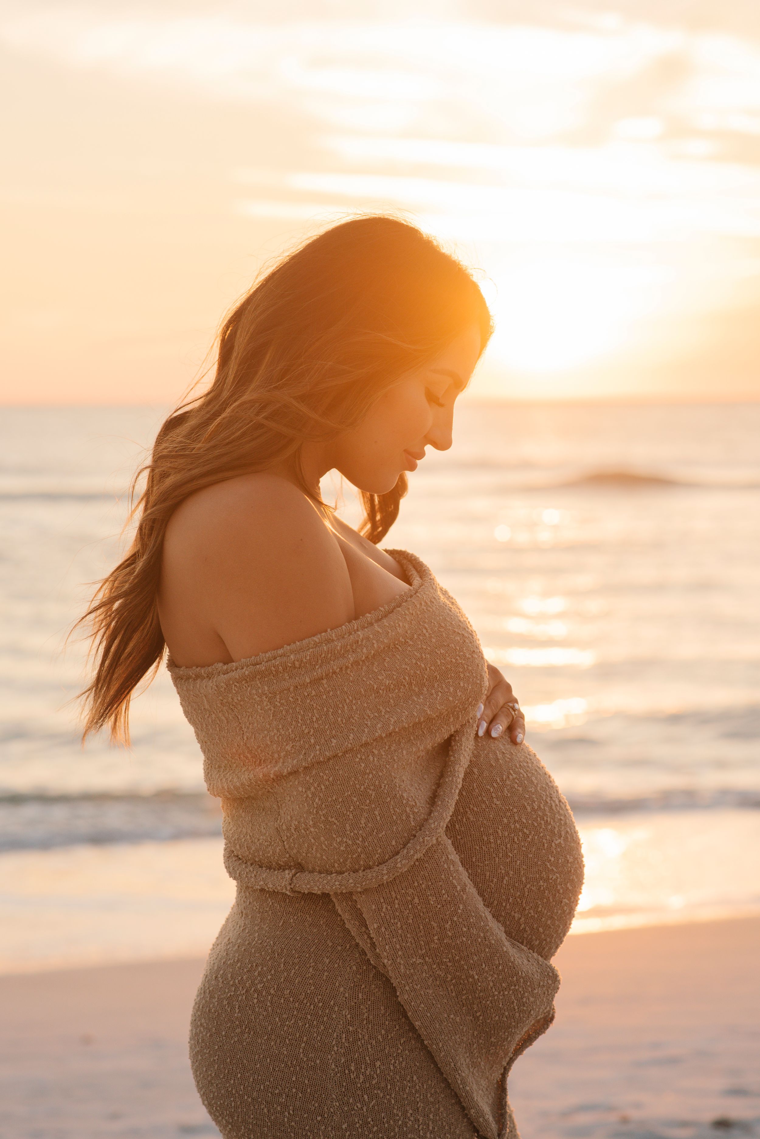 Silhouette of pregnant woman in off-shoulder dress gazing down during beach sunset.