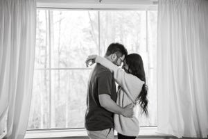 Intimate black and white photo of couple embracing by bright window with white curtains.