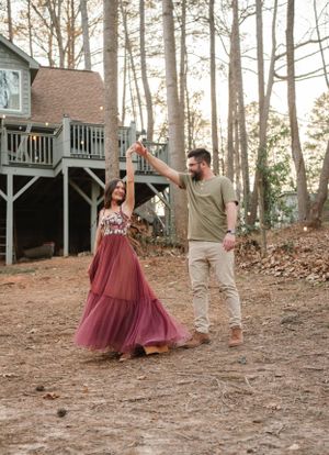 Dancing couple twirling near rustic cabin in wooded setting during sunset.
