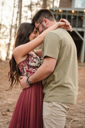 Romantic couple embraces outdoors during golden hour, with her burgundy dress flowing in the soft light.