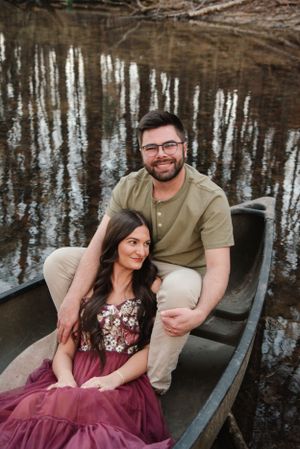 Couple poses together in a canoe on a calm lake with reflective water and trees in background.