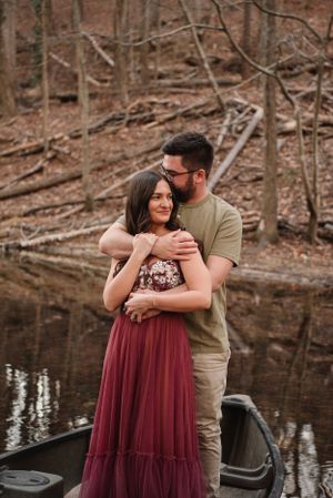 Couple embraces while standing in canoe on calm lake surrounded by winter trees.
