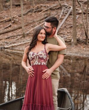 Couple poses together near lake with cabin in background, her wearing flowing burgundy dress.