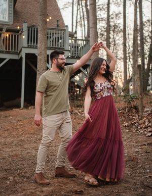 Dancing couple near cabin with burgundy dress twirling in forest setting.