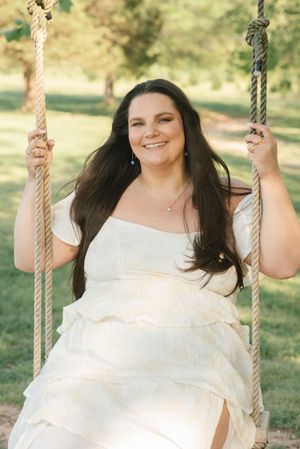 A person in a flowing white dress sits on a rope swing outdoors in a sunny park setting.
