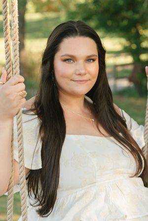 A person wearing a white lacy dress poses on a rope swing outdoors in a garden setting with trees in the background.