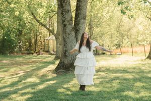 Person in flowing white tiered dress twirls gracefully under a large tree in a sun-dappled garden setting.
