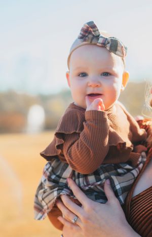Baby wearing plaid dress and brown sweater with matching bow headband in warm sunlight.
