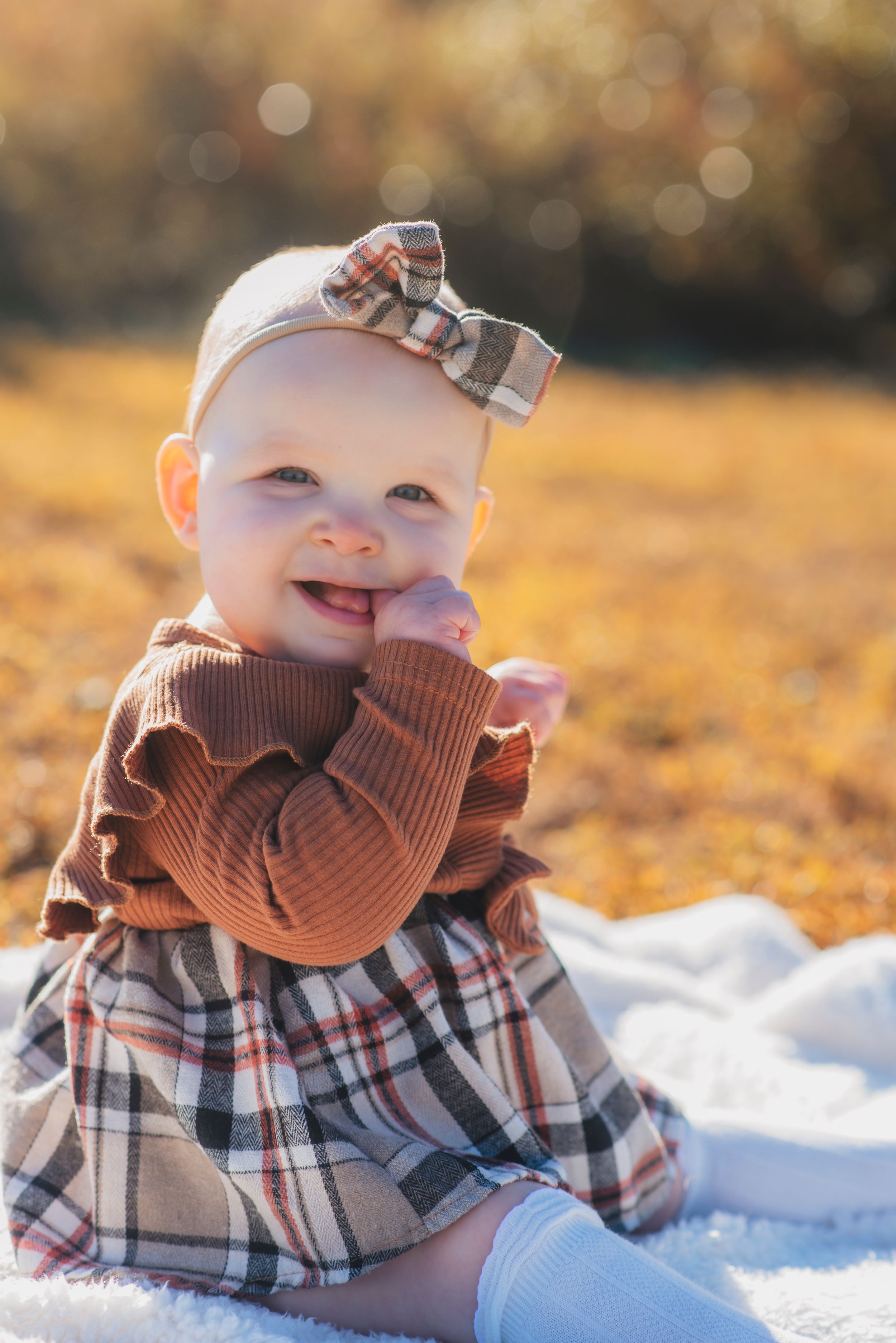 Smiling baby in plaid dress and bow headband sitting on white blanket in golden sunlight.