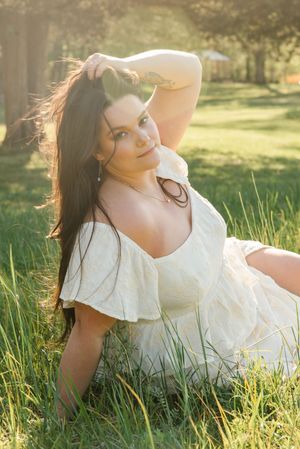 A soft sun-lit outdoor portrait of someone in a white flowing dress laying in golden summer grass.