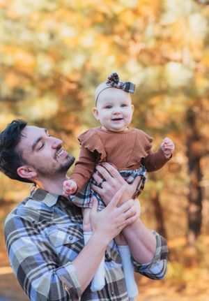 Playful outdoor moment with plaid-shirted parent lifting a smiling baby wearing a brown outfit against autumn trees.