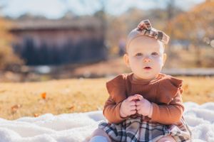 Baby in brown ruffled outfit sitting on white blanket outdoors with autumn scenery in background.