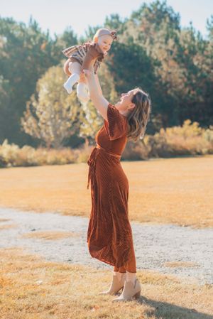 Parent lifting child high in the air while wearing long rust-colored dress against autumn background.