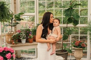 A tender moment shared between parent and baby in a sunlit greenhouse filled with pink flowers and lush green plants.