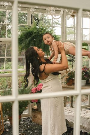 A tender moment shared between woman and baby in a sunlit greenhouse with ferns and pink flowers.