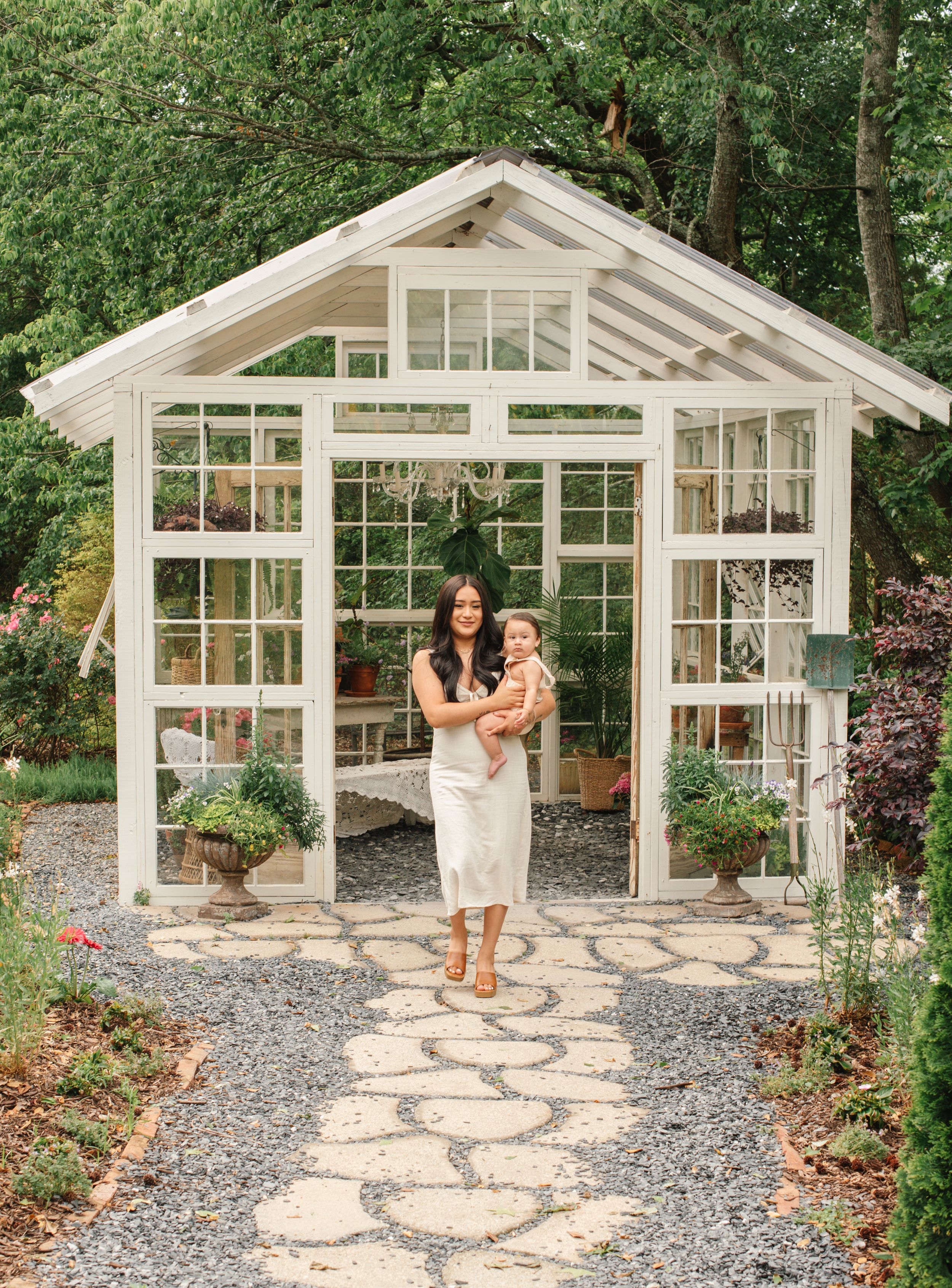 A charming white greenhouse with stone path features potted plants and garden decor in a wooded backyard setting.