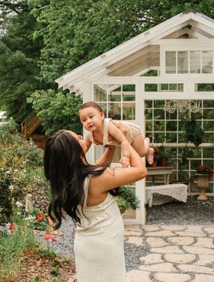 A tender moment between parent and baby takes place in front of a white greenhouse surrounded by lush gardens.