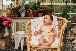 Baby sits in wicker chair surrounded by potted flowers and lace decor in sunny greenhouse setting.