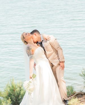 A couple shares a romantic kiss during their wedding ceremony on a scenic cliff overlooking the ocean at sunset.