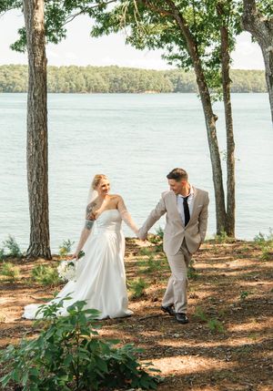 A romantic wedding couple strolls along a serene lakeside path beneath tall trees on a sunny afternoon.