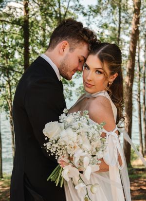 Romantic newlyweds embrace in a sun-dappled forest setting with a luxurious white floral bouquet and formal attire.