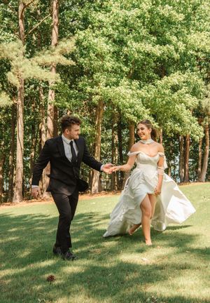 A couple in wedding attire runs playfully together on a sunny lawn surrounded by lush green trees.