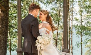 Romantic outdoor wedding scene with couple sharing intimate moment under wooden gazebo with lake backdrop and white bouquet.