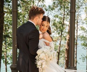 A romantic wedding moment captured at an outdoor gazebo surrounded by trees with the couple embracing and a white bouquet.