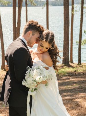 Newlyweds share intimate moment among tall trees by lakeside with bride holding white floral bouquet in sunlit forest.