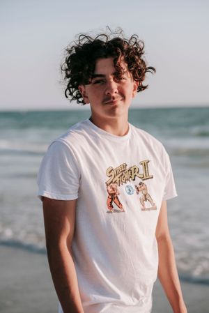 A person in a white graphic t-shirt smiling on a beach with curly dark hair and ocean waves in the background.