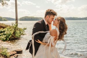A romantic couple in wedding attire embrace by a scenic lakeside during golden hour.