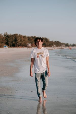 A casual beachside portrait of someone walking barefoot along the shoreline wearing jeans and a white t-shirt.